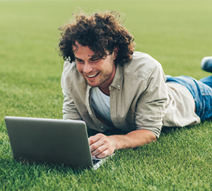 Young male laying on his stomach outside in the grass and smiling while working on his laptop.