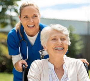 Caregiver in scrubs with a stethoscope around her neck pushing a patient in a wheelchair.